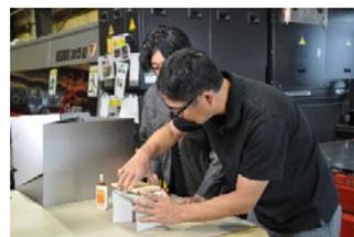 A man in a black t-shirt and safety glasses working with a tool on a workbench in a workshop.